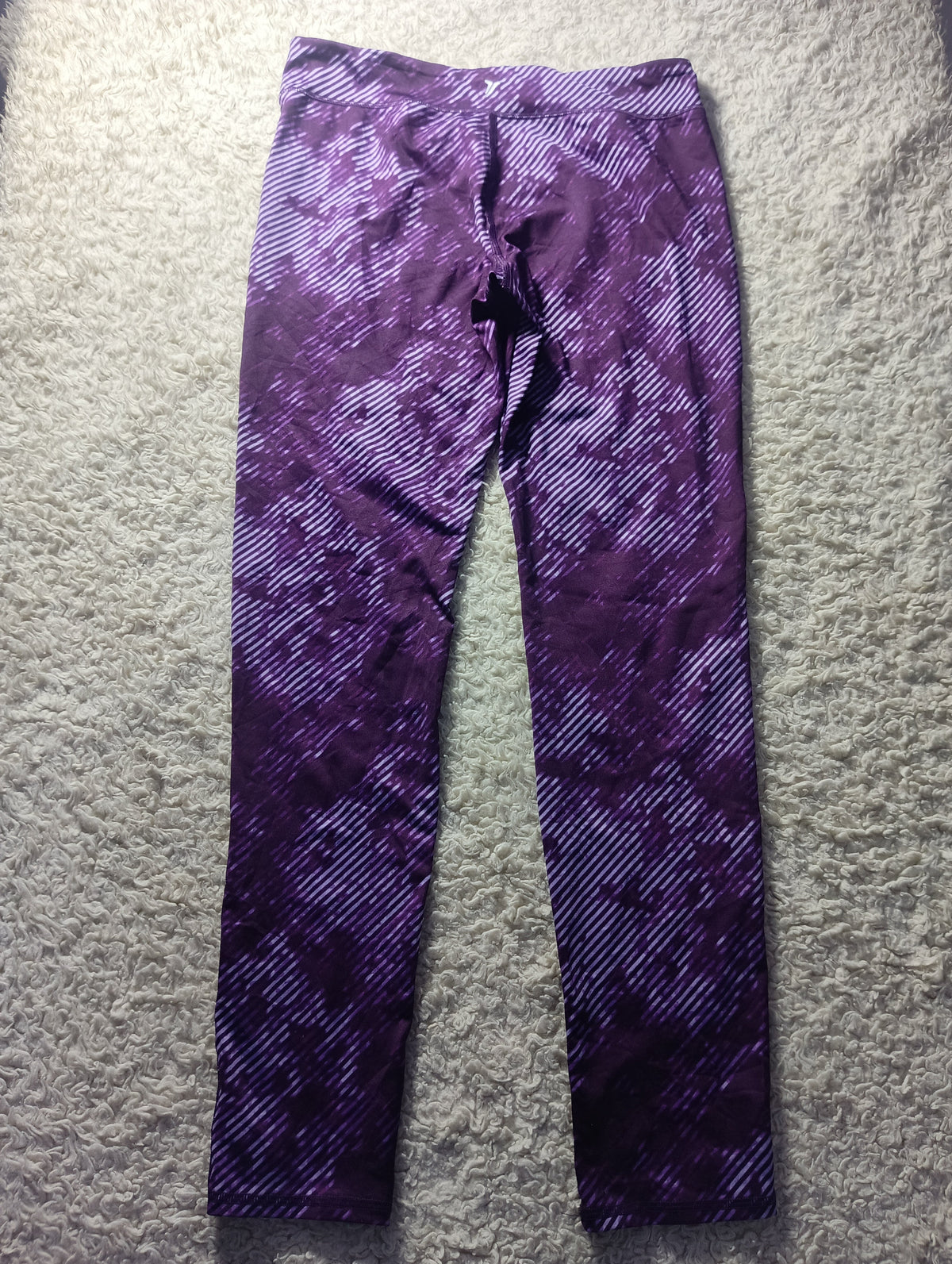 Purple patterned athletic leggings laid flat on a gray textured carpet. The leggings feature a diagonal striped pattern that fades into a solid dark purple at the lower legs.