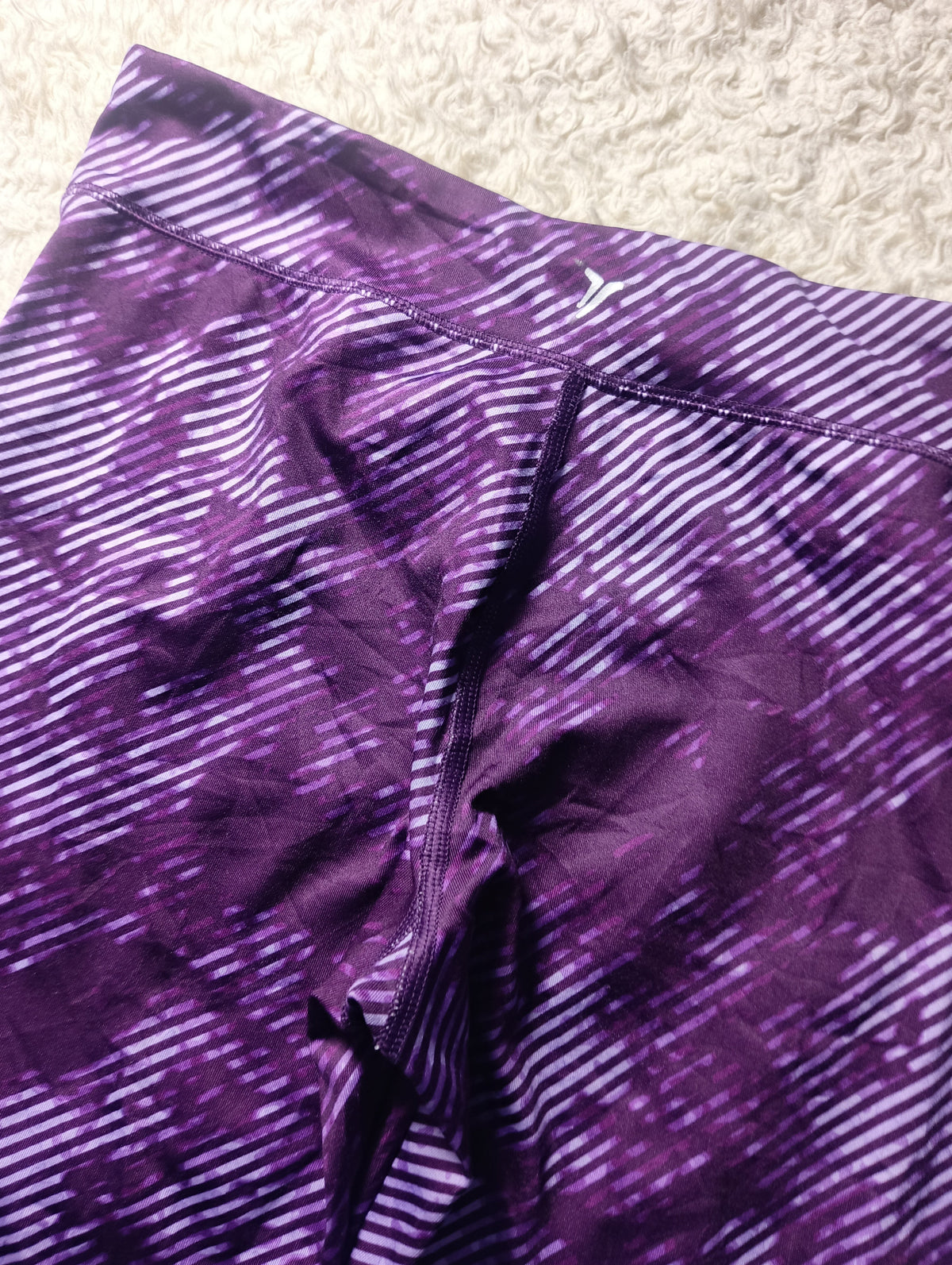 Purple athletic leggings with a diagonal stripe pattern and a small white Y-Brand logo near the waistband, laid flat on a textured beige carpet.