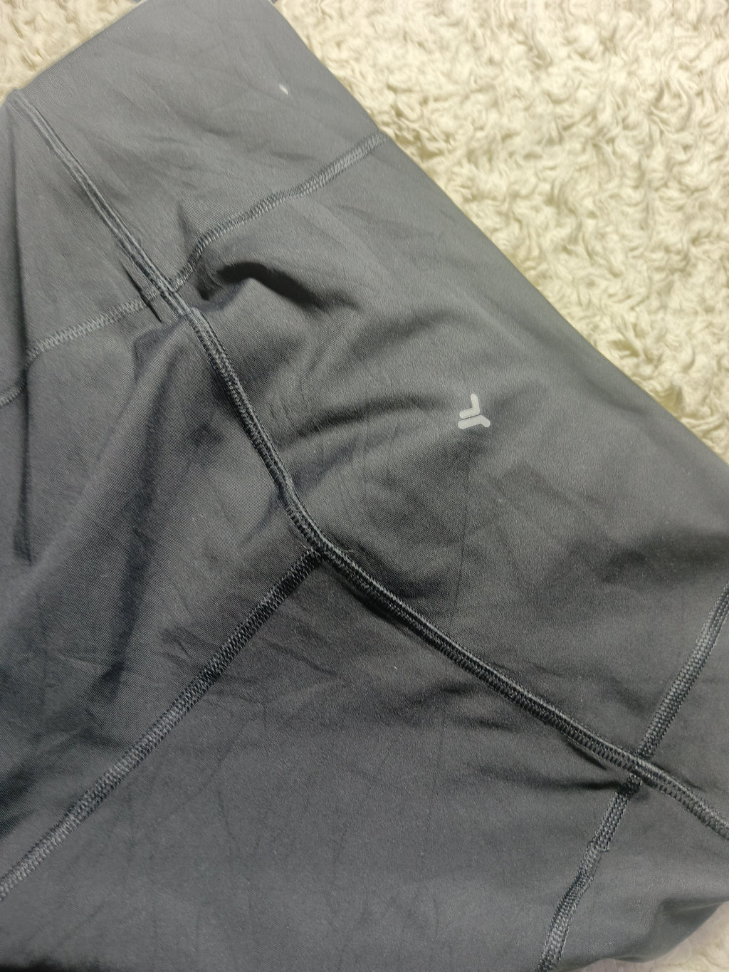 A close-up of dark gray athletic leggings laid flat on a beige textured rug, showing the fabric texture, seams, and a small white embroidered logo.