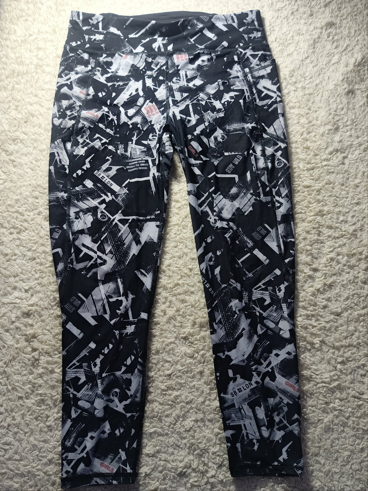 Black and white abstract patterned leggings laid flat on a beige carpet. The image has a camera watermark overlay at the bottom.
