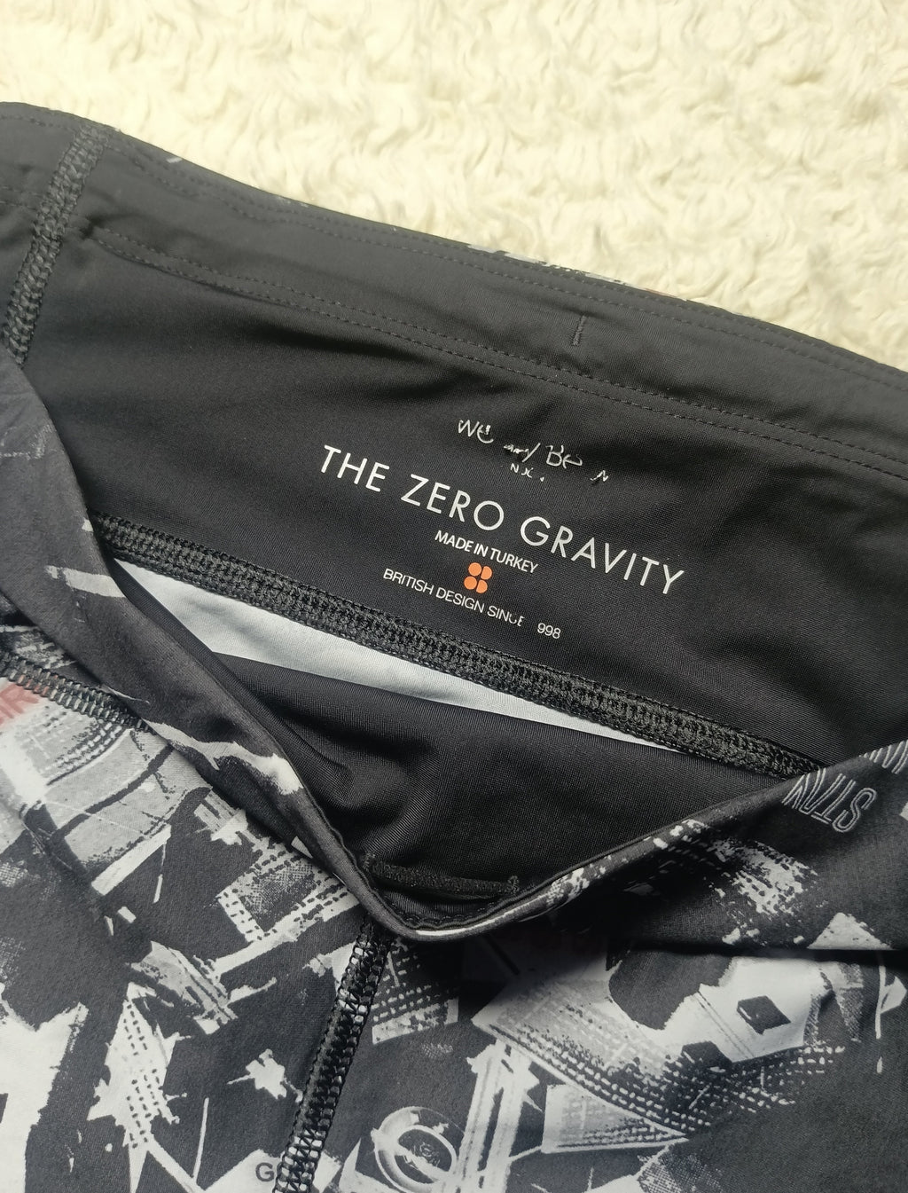 Close-up of a black athletic garment showing the inner neck label with 'THE ZERO GRAVITY' branding, alongside a black-and-white abstract print on the fabric, photographed on a textured cream surface.