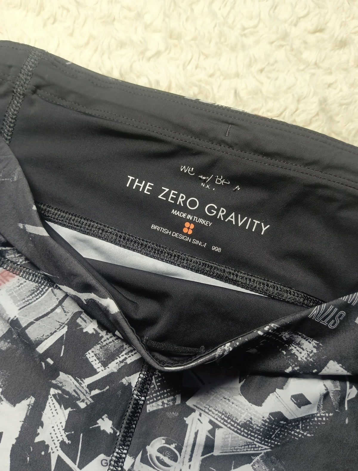 Close-up of a black athletic garment showing the inner neck label with 'THE ZERO GRAVITY' branding, alongside a black-and-white abstract print on the fabric, photographed on a textured cream surface.