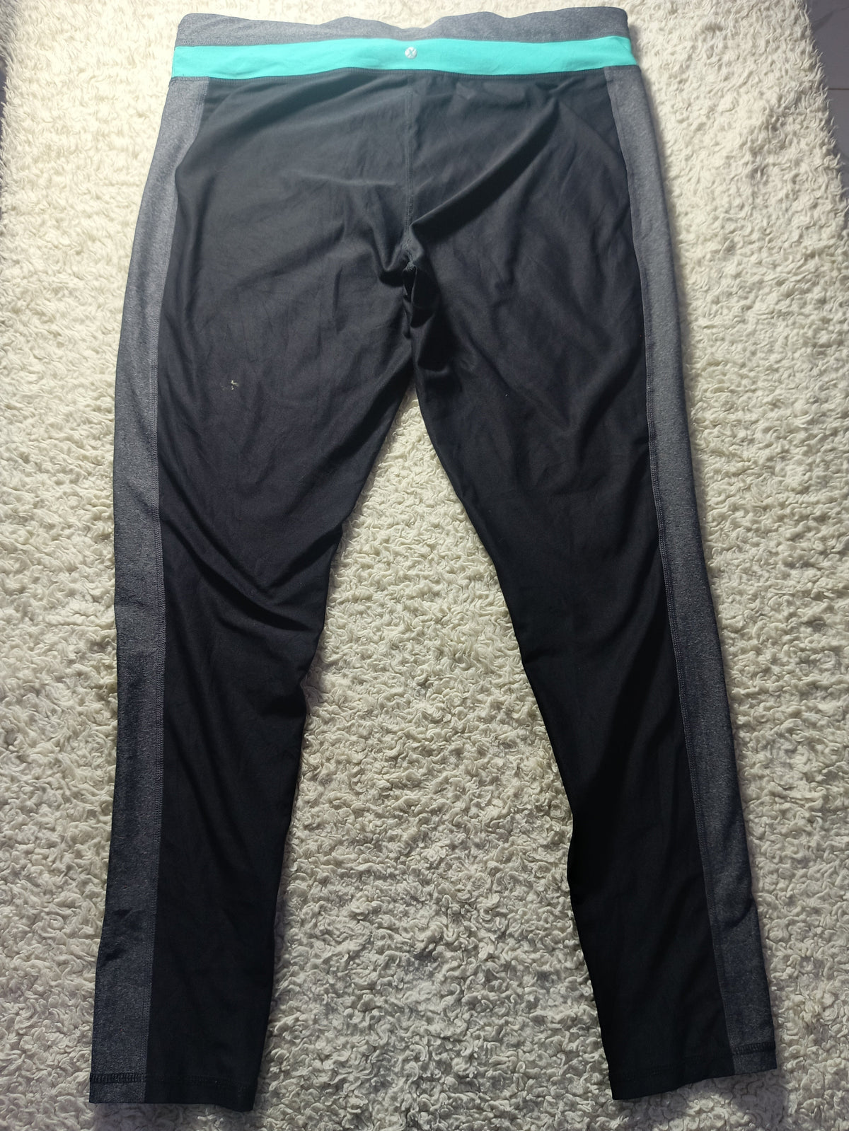 Black athletic leggings with a bright teal waistband and dark gray side panels, laid flat on a textured gray carpet. A small white circular logo is visible at the center of the waistband.