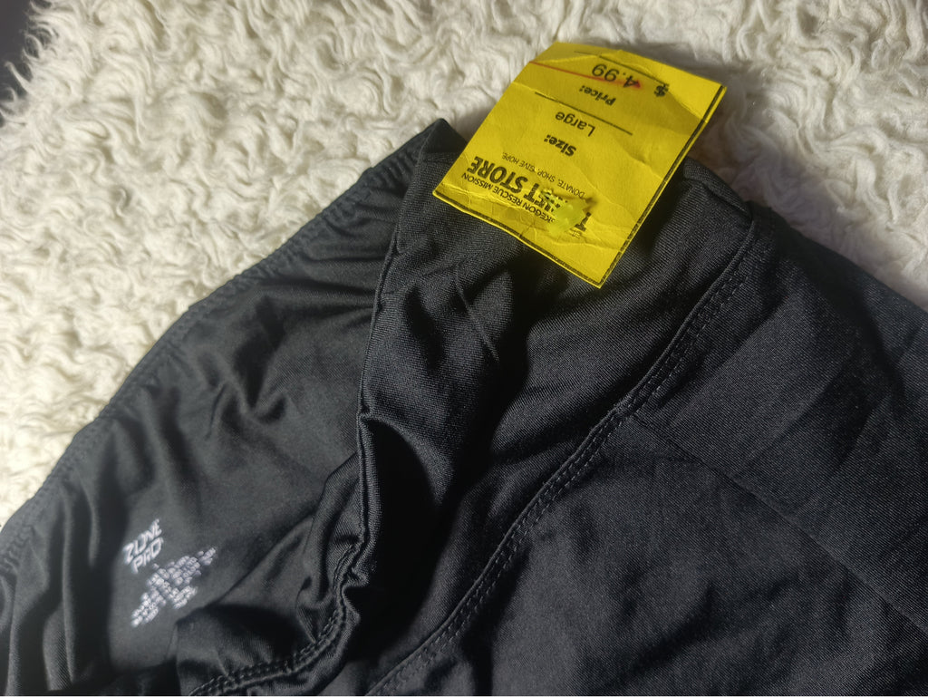 A close-up of black athletic shorts laid flat on a white shag rug, showing the elastic waistband, inner leg construction, and a bright yellow size tag.