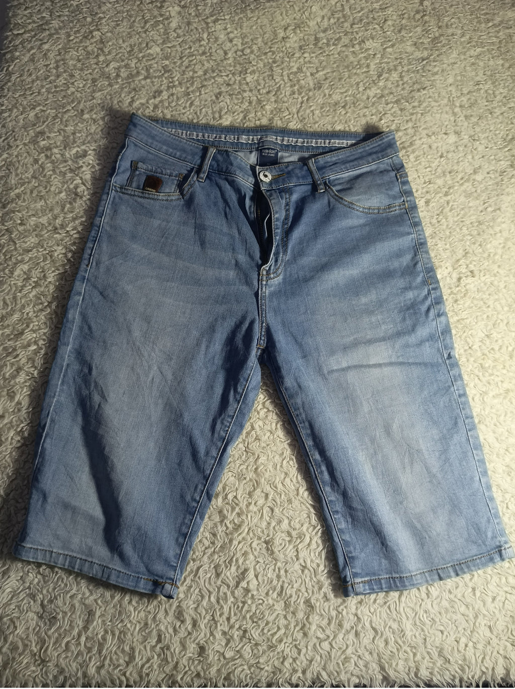 Light blue denim capri jeans laid flat on a textured gray carpet, showing the front waistband, belt loops, and front pockets.
