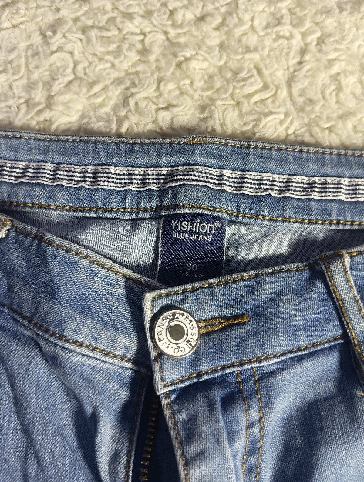 Close-up of the waistband and button area of blue denim jeans, showing a visible Xishion brand label and a silver button on a textured cream background.
