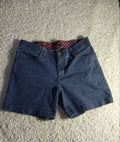 A pair of dark blue denim shorts laid flat on a textured beige carpet, showing a red and black patterned lining inside the waistband.