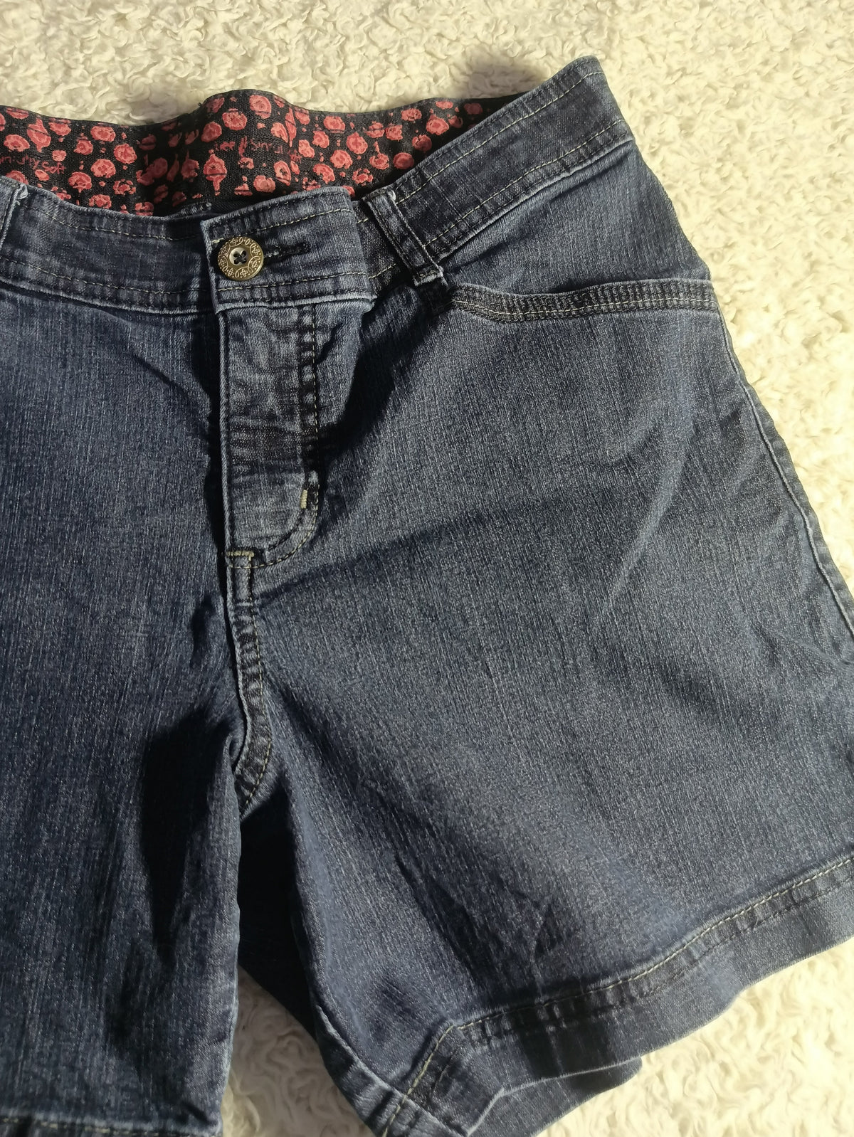 A pair of dark wash denim shorts laid flat on a light beige textured surface, showing the front pocket, button closure, and a patterned red and black lining inside the waistband.