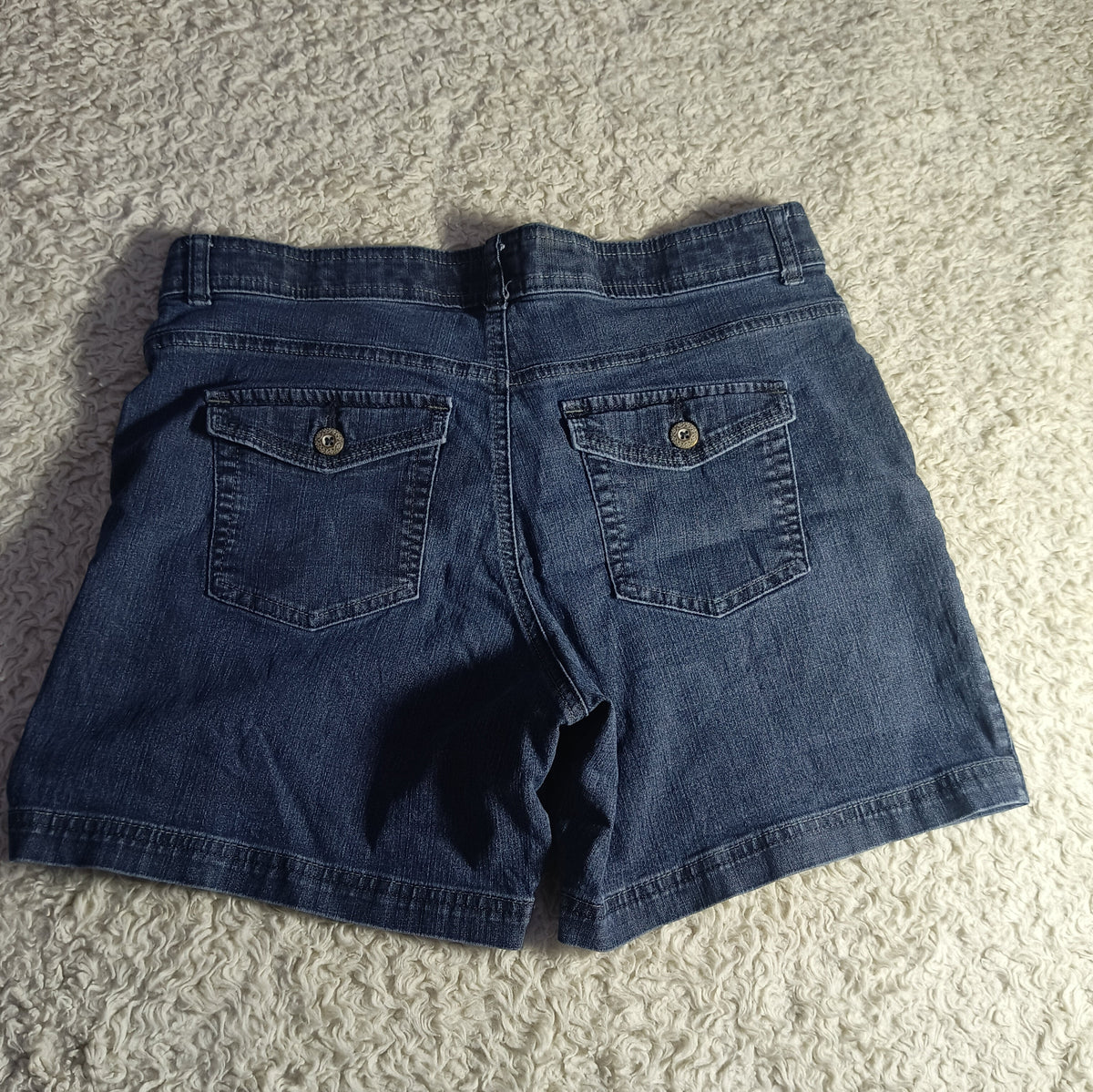 Back view of dark blue denim shorts laid flat on a textured beige carpet, showing two buttoned back pockets.