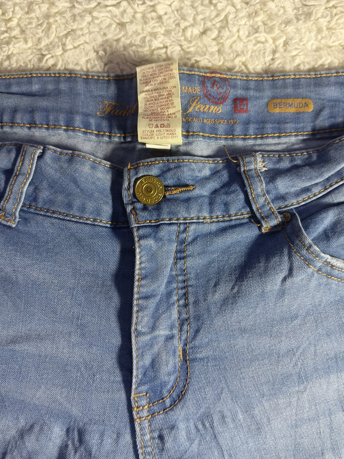 Close-up of a pair of light blue denim jeans laid flat on a textured cream fabric background, showing the waistband, button closure, and inner label details.