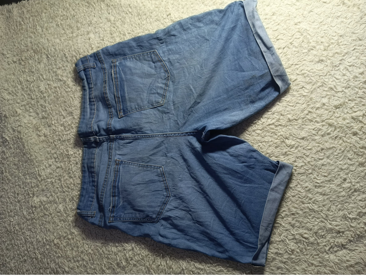 A pair of blue denim shorts laid flat on a textured beige carpet, photographed from above. The shorts have a faded medium-wash blue color, visible back pockets, and rolled cuffs.