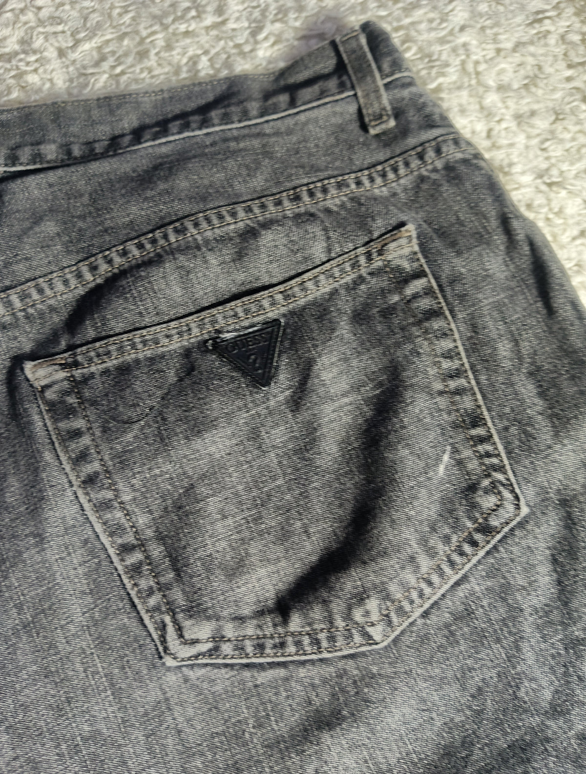 Close-up of the back pocket of gray washed denim jeans, showing the triangular Guess logo patch on the pocket and the waistband stitching, photographed on a textured gray surface.