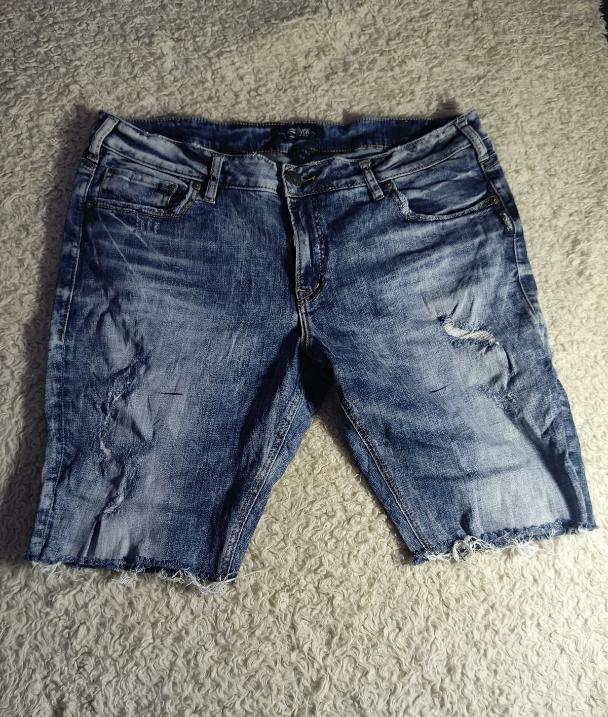 A pair of distressed blue denim shorts with frayed hems, laid flat on a textured gray carpet.