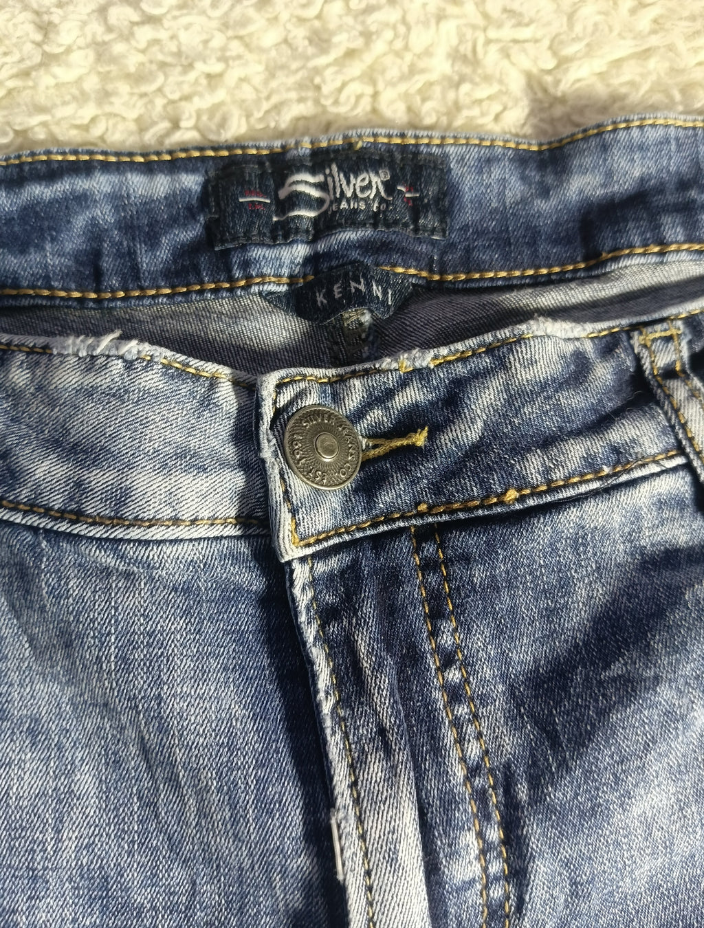 Close-up of a pair of blue denim jeans showing the waistband, button closure, and inner label details, laid flat on a textured cream surface.