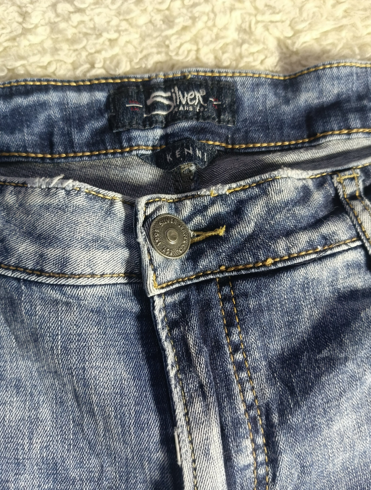 Close-up of a pair of blue denim jeans showing the waistband, button closure, and inner label details, laid flat on a textured cream surface.