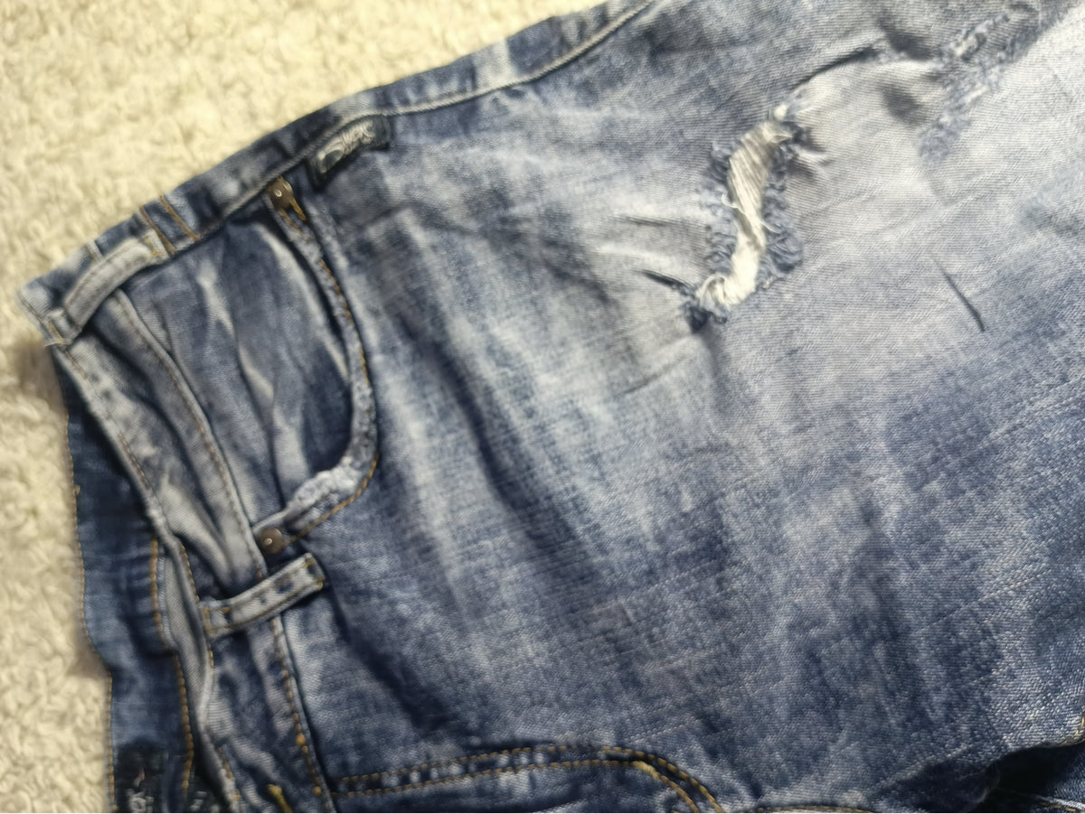 Close-up of a pair of blue denim shorts with a distressed rip and whiskering, laid flat on a beige carpet. A white text bar is at the bottom with camera settings and POVA & Neo branding.