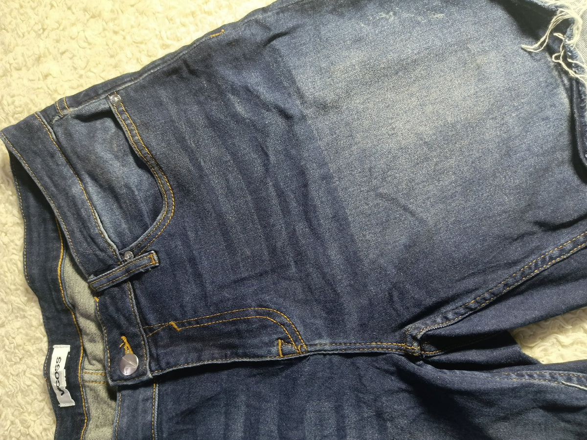 A close-up of a pair of dark blue denim jeans laid flat on a light beige textured surface, showing the waistband, belt loops, pocket, and stitching details.