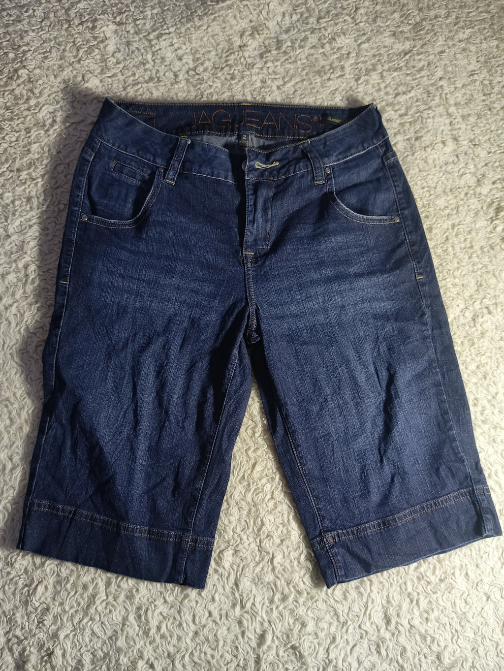 A pair of dark blue denim capri pants laid flat on a textured gray carpet. The pants feature a classic five-pocket design, belt loops, and a dark wash.