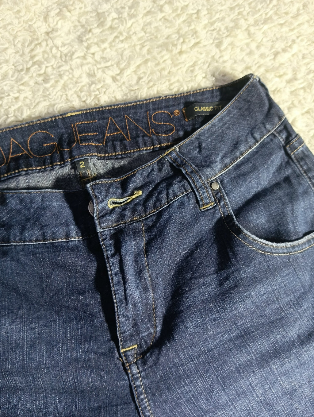 A close-up of a pair of dark blue denim jeans laid flat on a textured cream fabric background, showing the waistband, belt loops, button, and front pocket.
