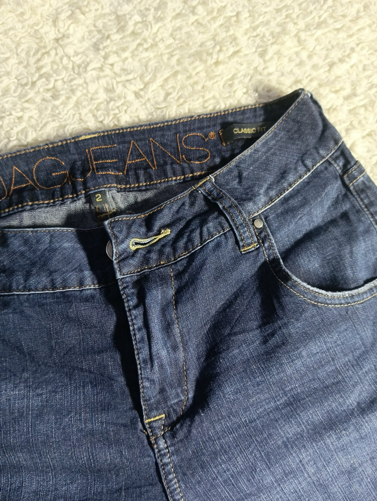 A close-up of a pair of dark blue denim jeans laid flat on a textured cream fabric background, showing the waistband, belt loops, button, and front pocket.