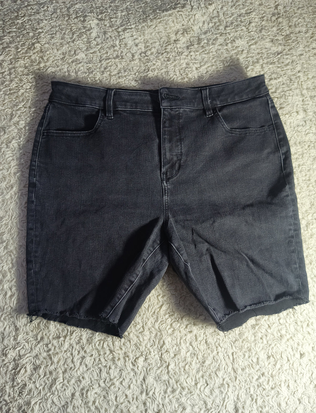 Dark charcoal gray denim shorts laid flat on a textured beige carpet. The photo is taken from above and includes a camera watermark at the bottom.