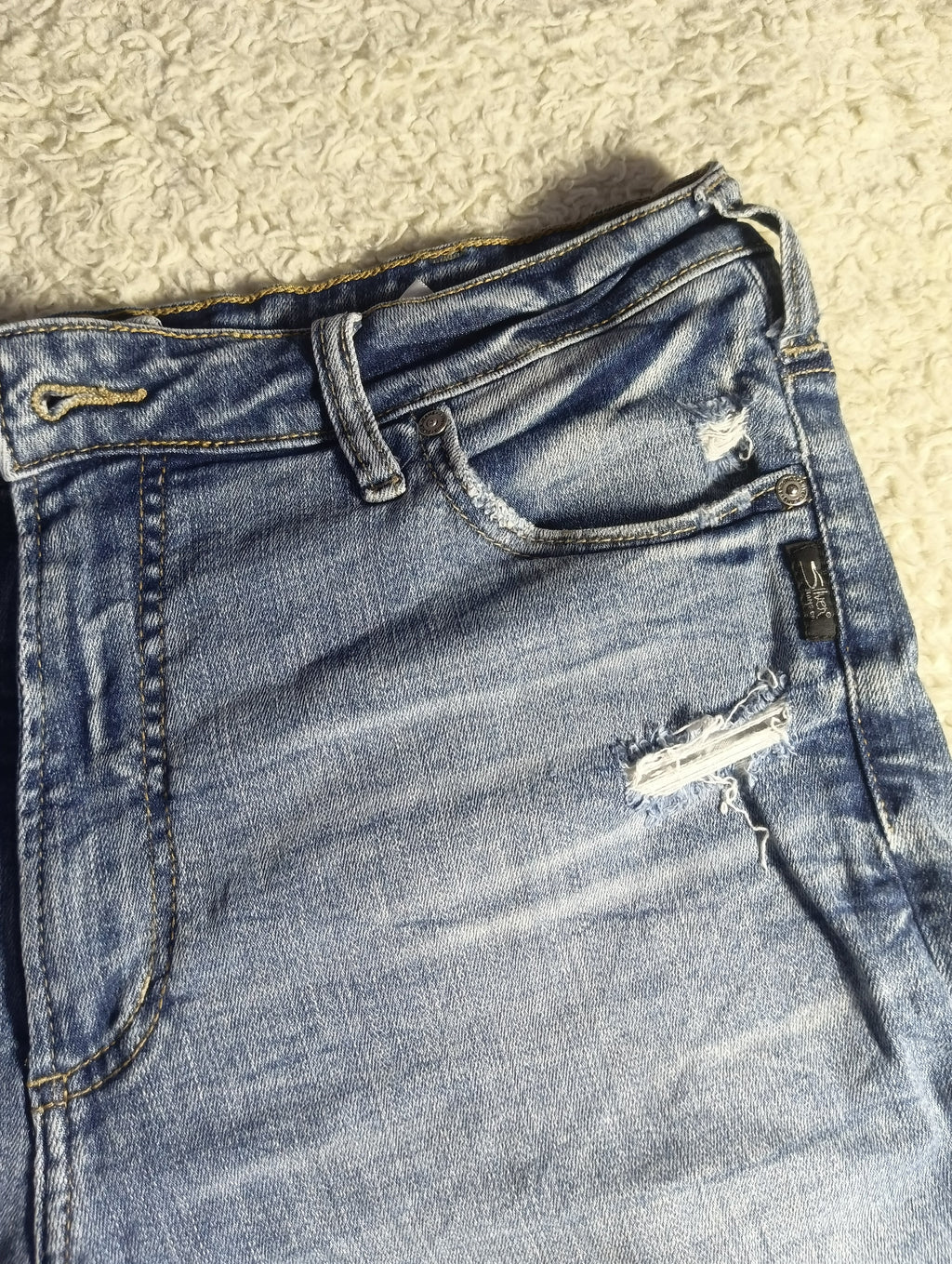 Close-up of a pair of light-wash blue denim jeans laid flat on a textured beige carpet, showing the front pocket, belt loops, and distressed rips on the thigh.