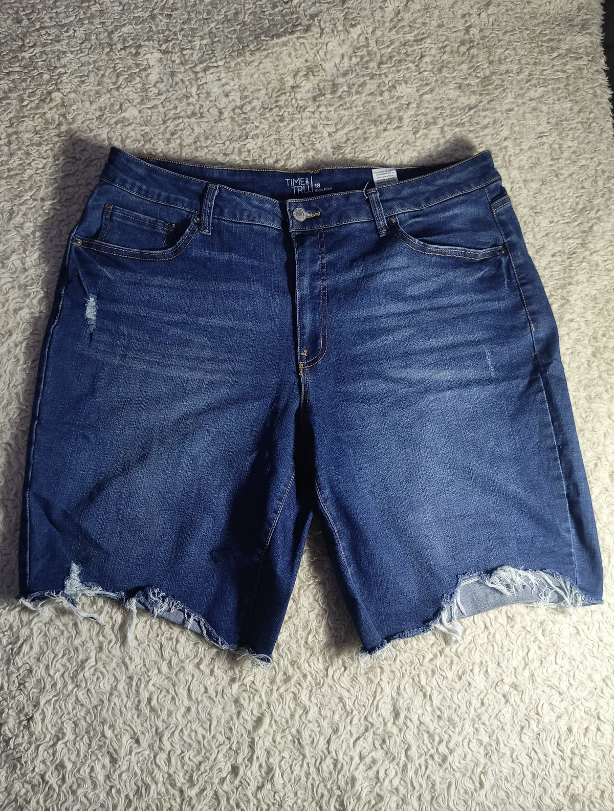 A pair of blue denim shorts with a distressed wash and raw frayed hems, laid flat on a textured gray carpet.
