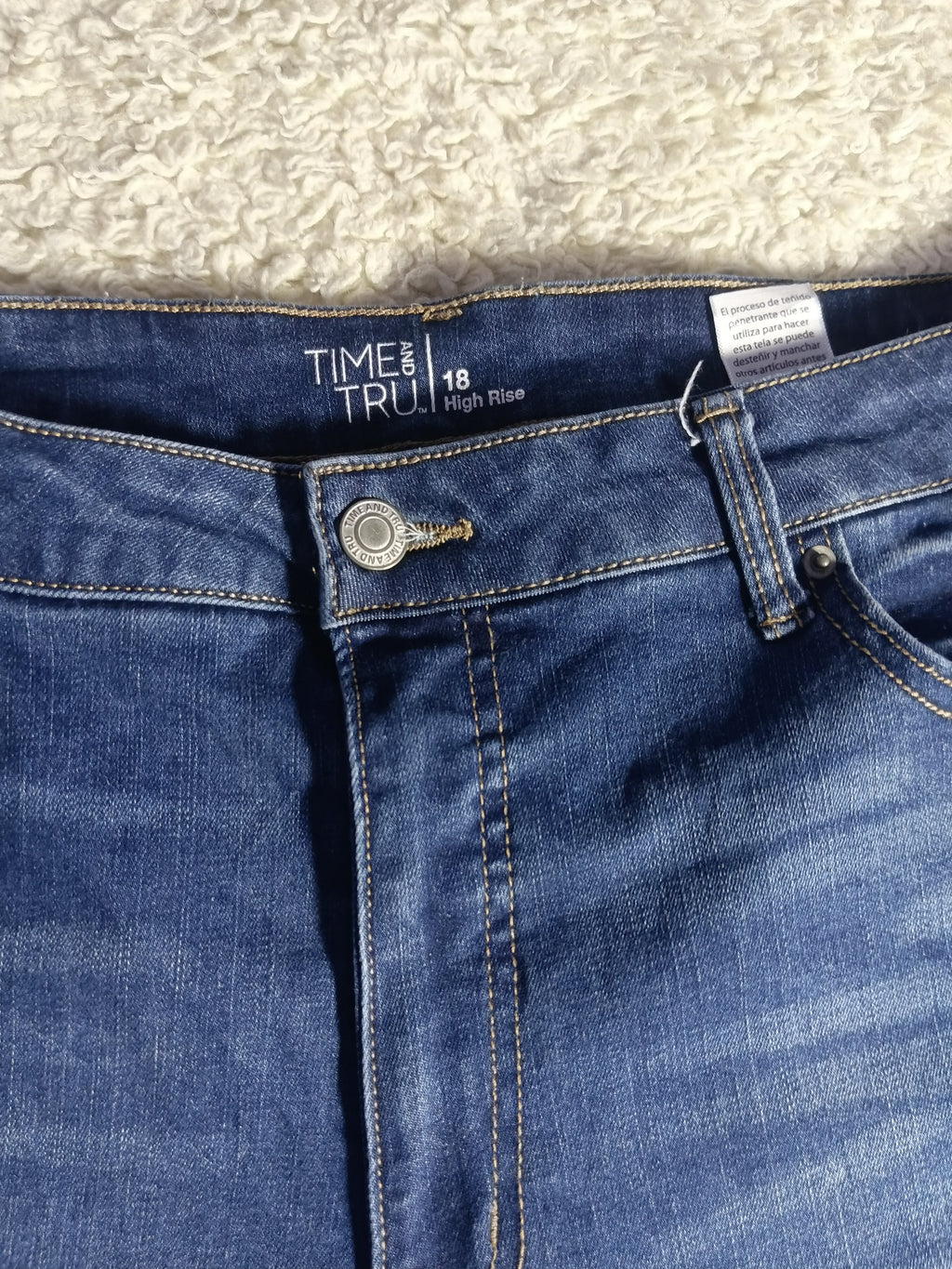 Close-up of the waistband of blue denim jeans, showing the button, belt loops, front pocket, and inner label that reads TIME & TRUL 18 High Rise.