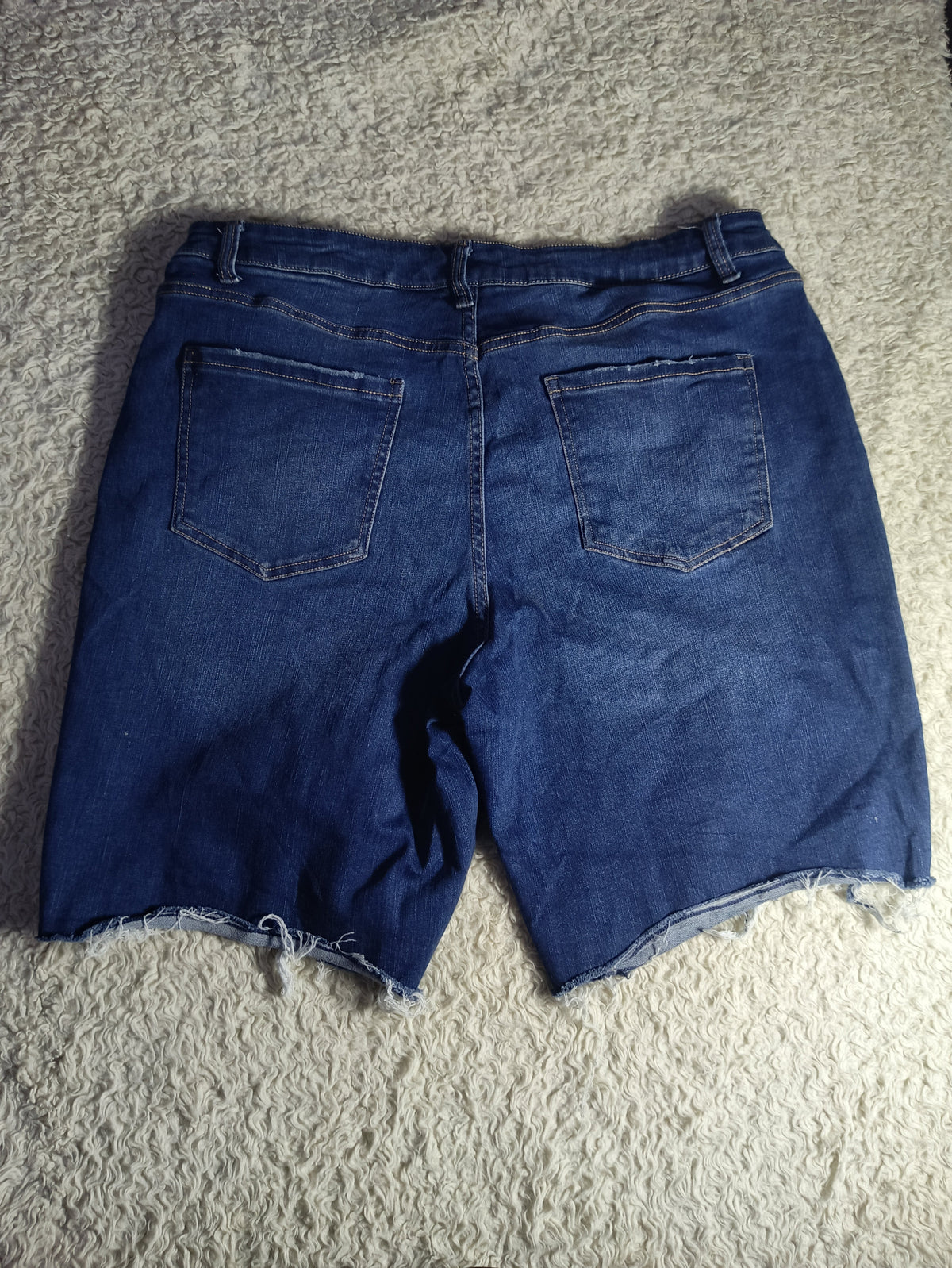 Back view of dark blue denim shorts with a raw, frayed hem, laid flat on a textured gray carpet.