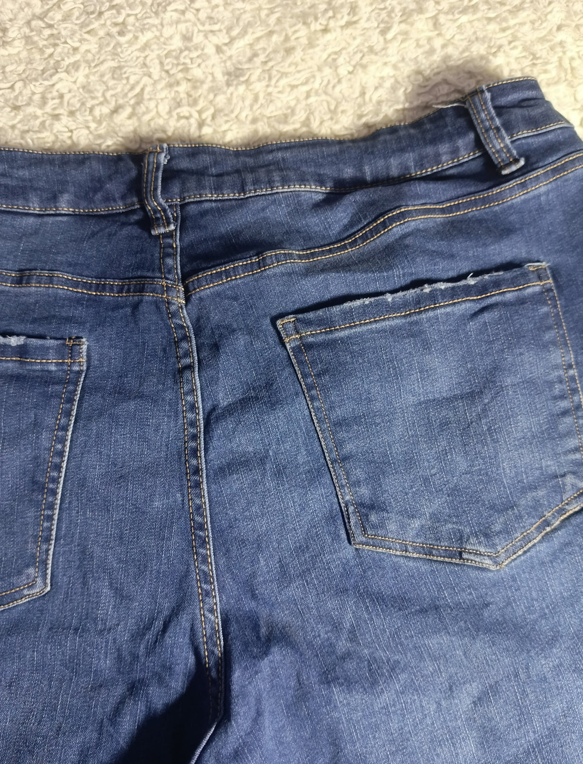 Close-up back view of blue denim jeans showing the waistband, belt loops, back pockets, and stitching, photographed on a textured light beige surface.