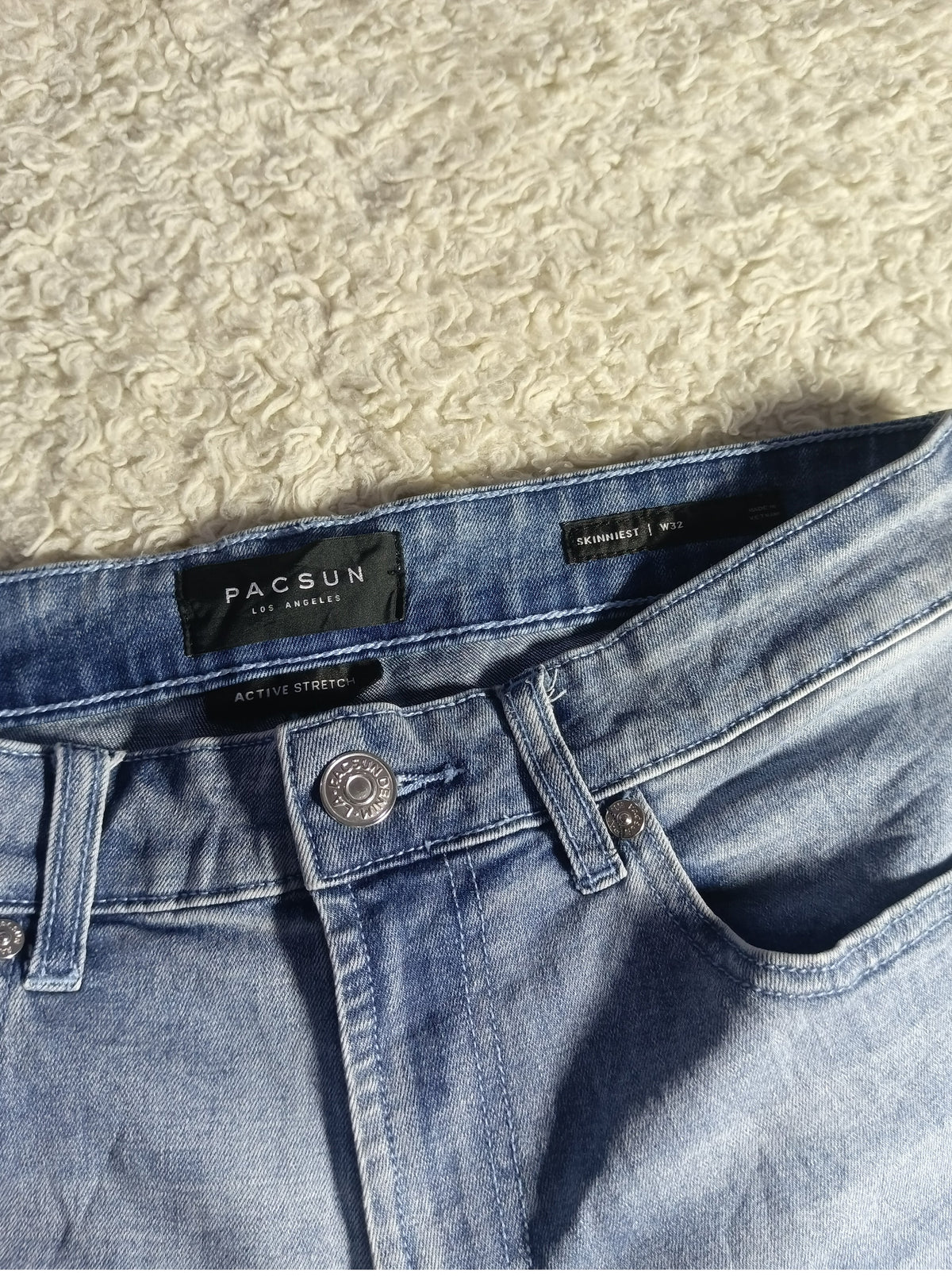 A close-up of light blue denim jeans laid flat on a textured beige carpet, showing the waistband, black PACSUN brand label, button, and pocket.