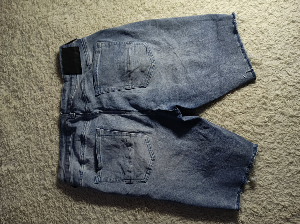 A pair of faded blue denim shorts laid flat on a textured beige carpet, viewed from above. The shorts feature a classic five-pocket design and a raw, frayed hem.