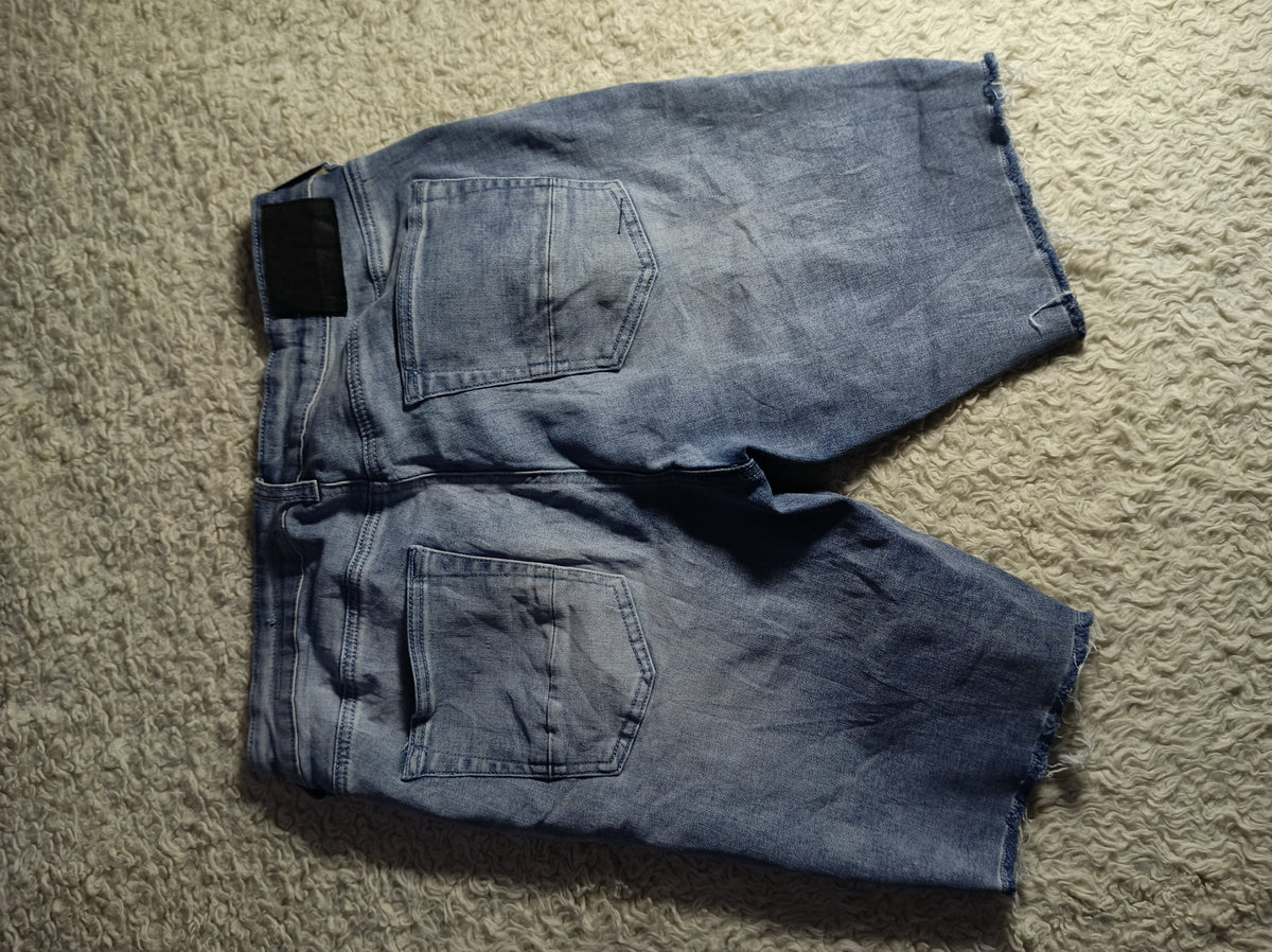 A pair of faded blue denim shorts laid flat on a textured beige carpet, viewed from above. The shorts feature a classic five-pocket design and a raw, frayed hem.