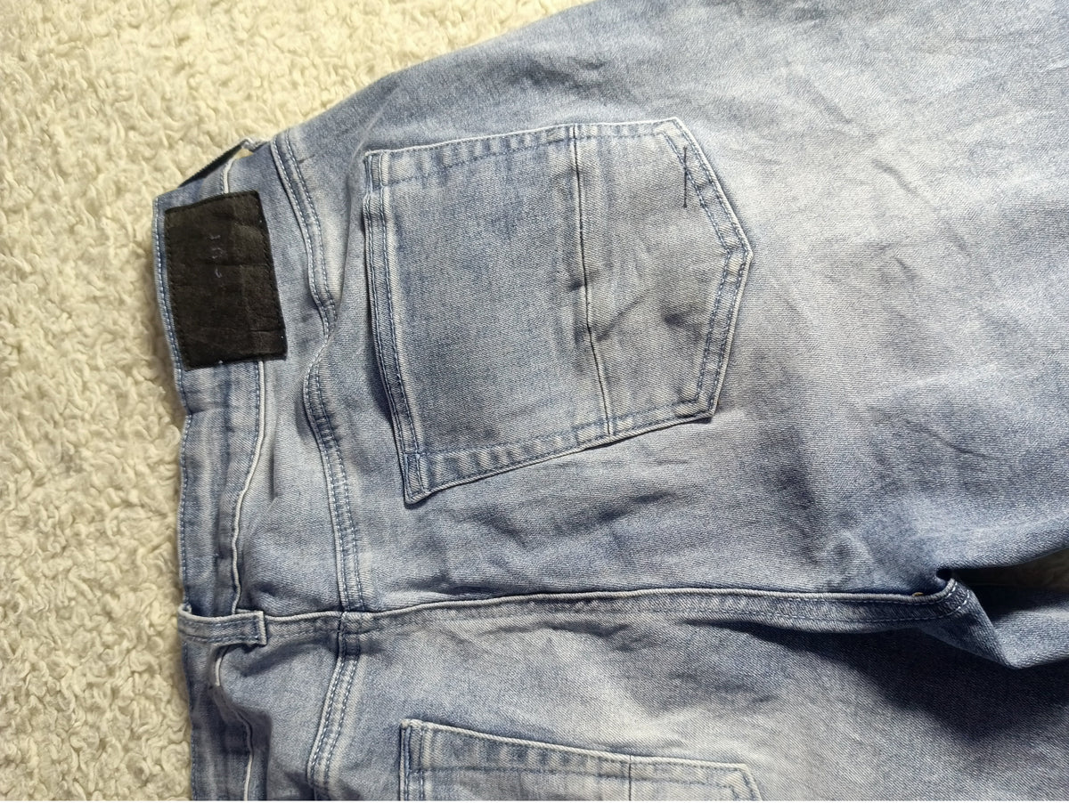 A close-up of the back waistband and pocket area of light-wash blue denim jeans laid flat on a textured beige carpet. A black fabric tag is visible near the back waistband.