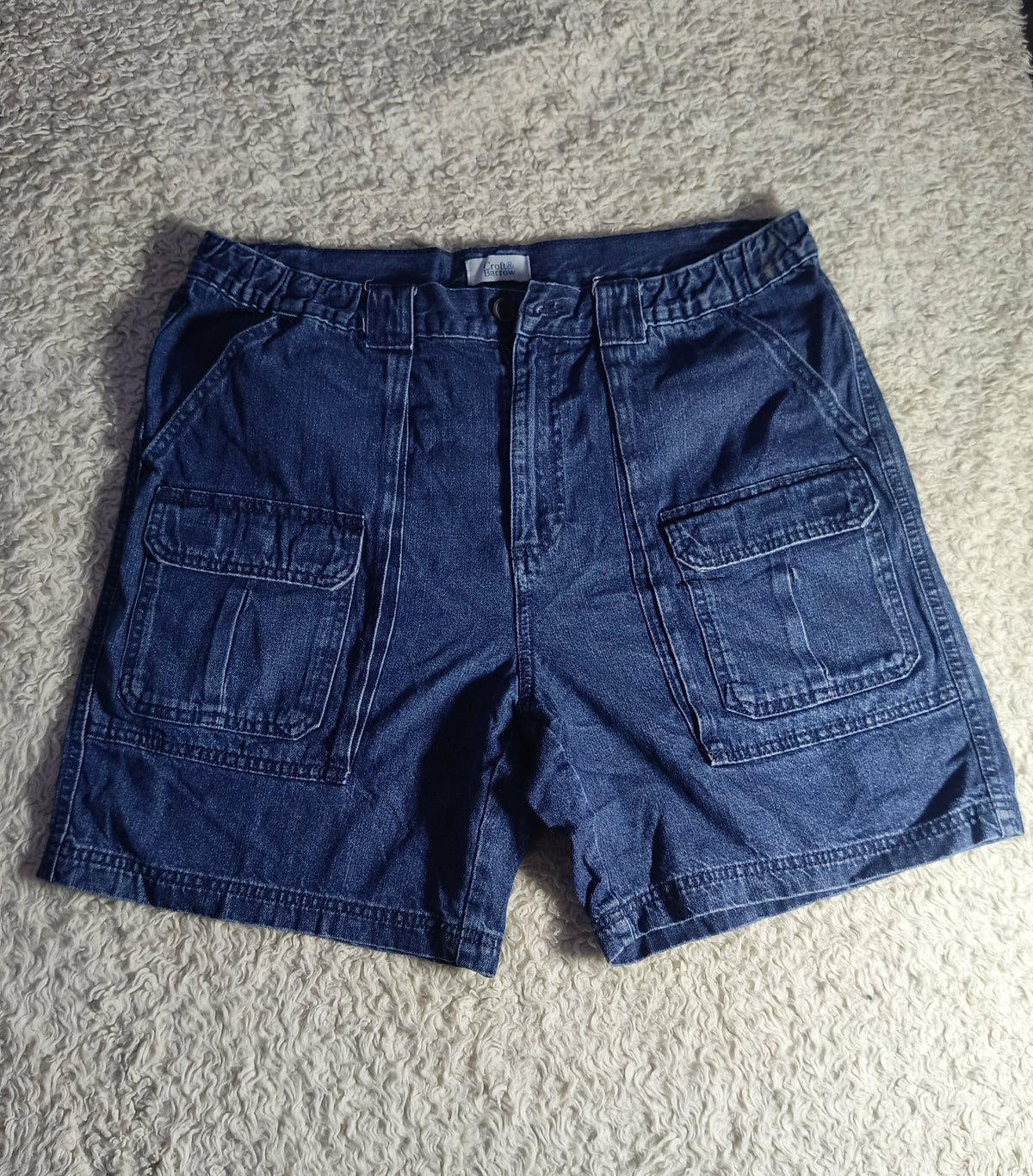 A pair of dark blue denim cargo shorts laid flat on a textured gray carpet. The shorts feature a button closure, belt loops, and two large front cargo pockets.