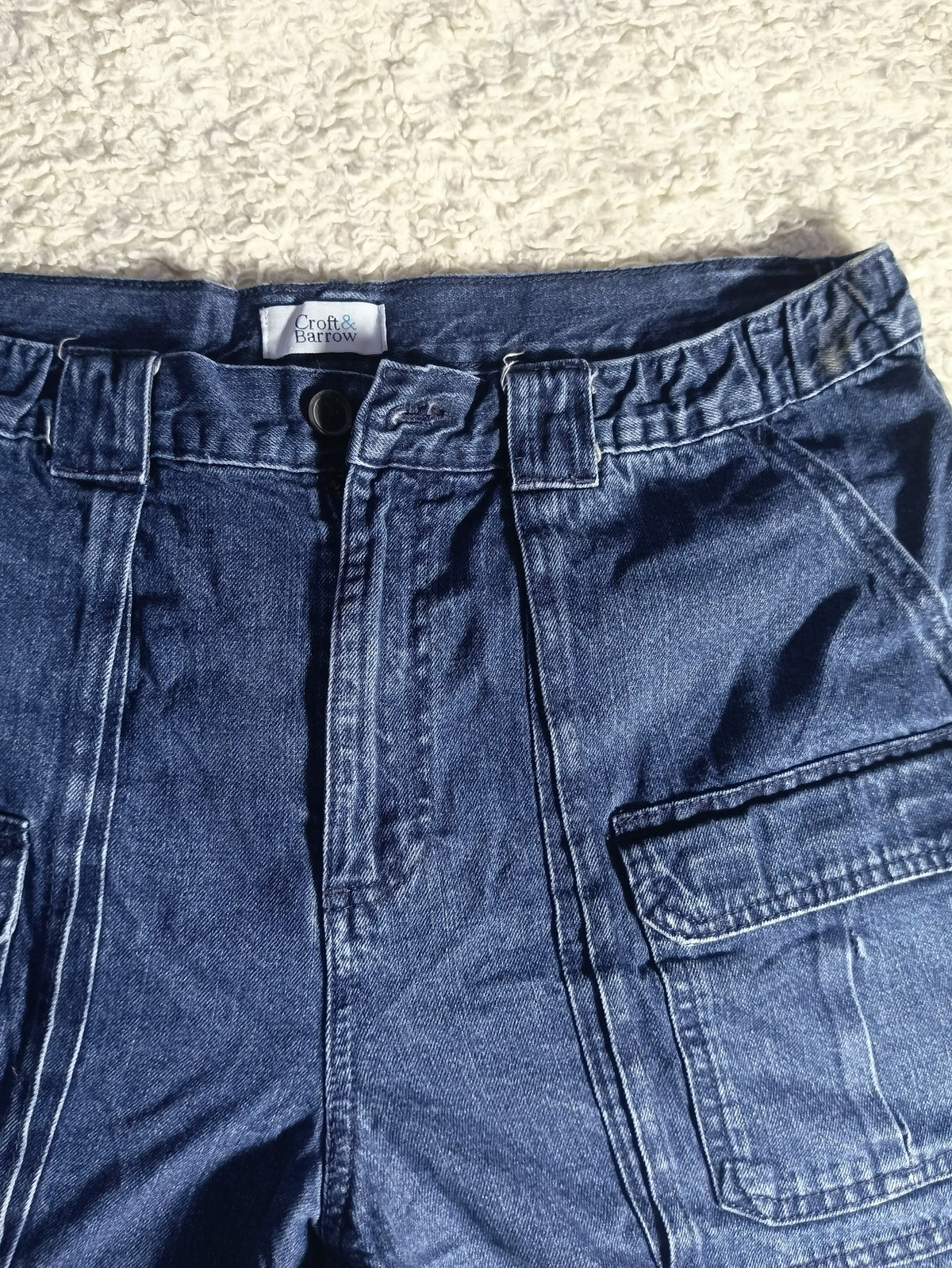A close-up of a pair of blue denim shorts laid flat on a textured beige carpet. The photo shows the waistband, front fly, belt loops, and a side cargo pocket.