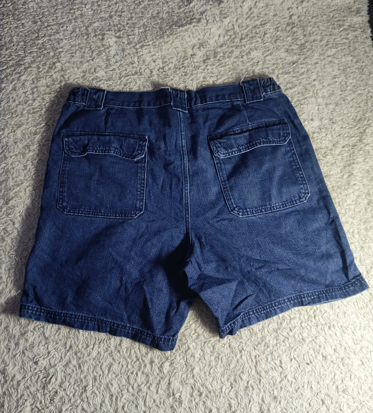 A pair of dark blue denim shorts shown from the back, laid flat on a textured gray carpet. The photo has a camera watermark overlay at the bottom.