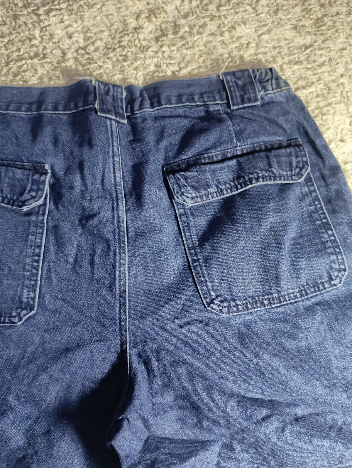Close-up of the back waistband and right rear pocket of blue denim jeans laid flat on a textured beige carpet. A dark shadow falls across the lower left portion of the denim.
