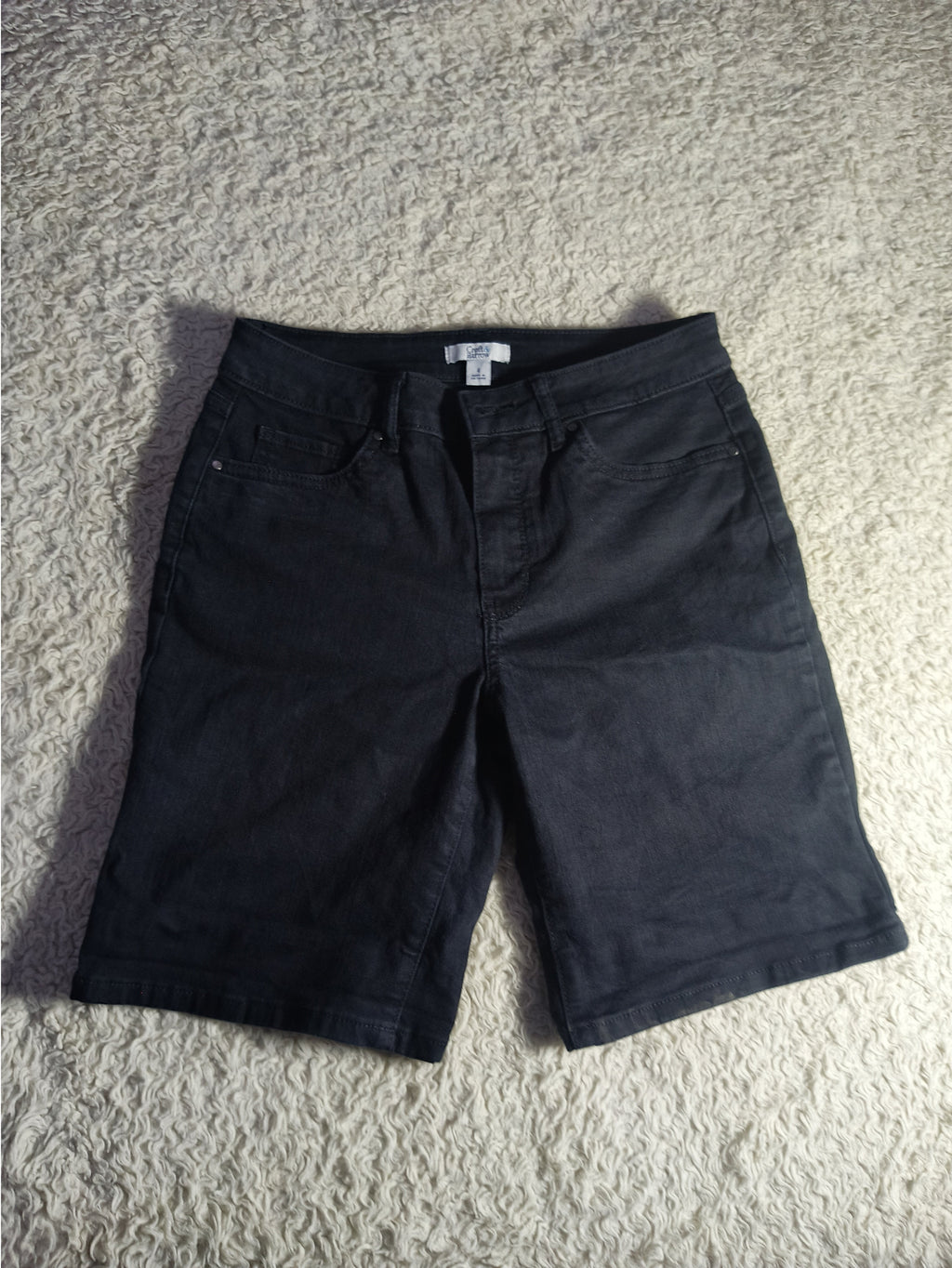 Black denim shorts laid flat on a textured beige carpet, with a visible waistband and front pocket.