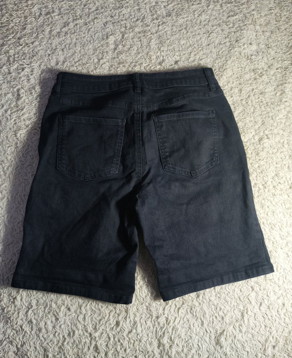 Black denim shorts viewed from the back, laid flat on a textured beige carpet.