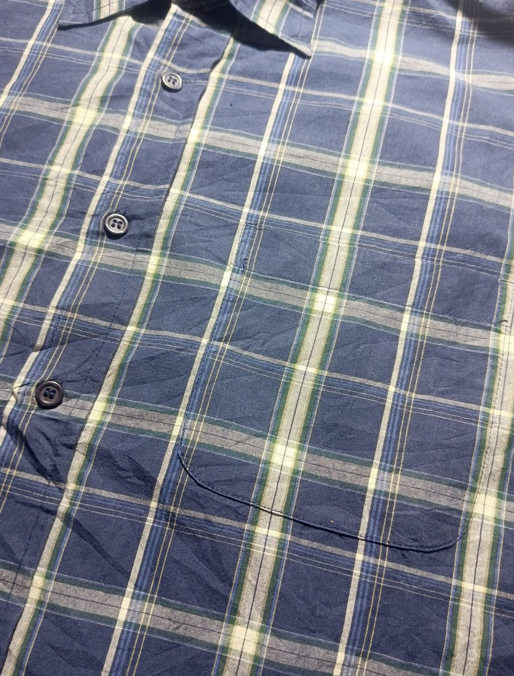 A close-up of a blue plaid button-up shirt fabric, showing the pattern details and dark buttons.