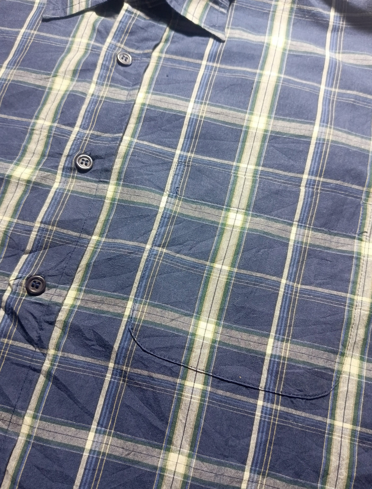 A close-up of a blue plaid button-up shirt fabric, showing the pattern details and dark buttons.
