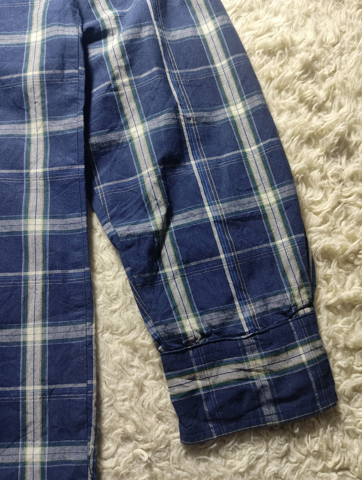 A close-up of a blue plaid shirt or pajama fabric laid on a cream shaggy surface, showing the cuff and part of the body of the garment. Small text at the bottom reads POVA & Neo and 23mm f/1.