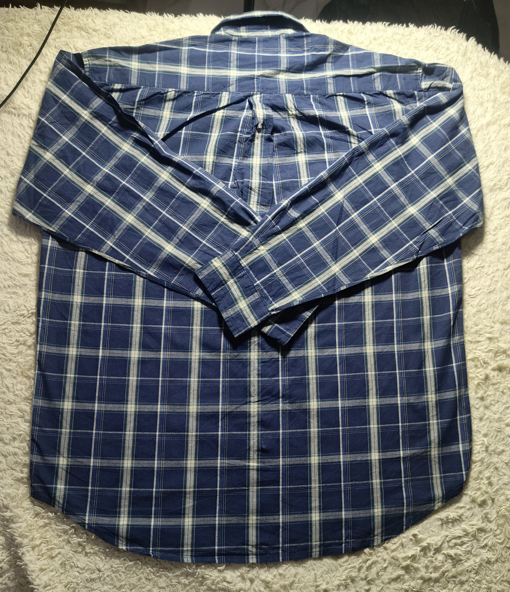 A blue and white plaid long-sleeve button-up shirt laid flat on a white textured blanket, with the collar standing up. The photo has a camera watermark at the bottom.