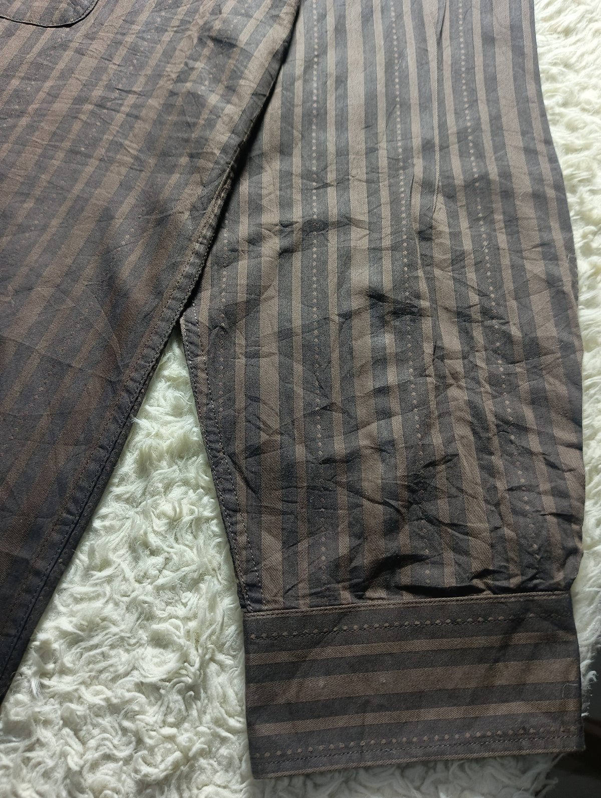 A close-up of a striped fabric garment laid flat on a white textured rug. The fabric has vertical and horizontal stripes in dark brown and gray tones, with a visible cuff showing horizontal stripes.