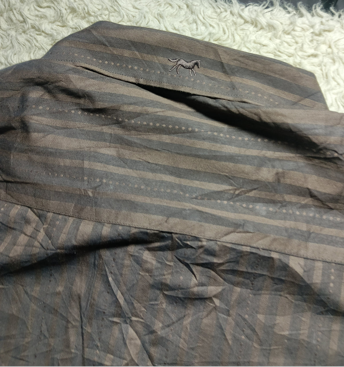 A close-up of a dark brown and black striped fabric garment laid out on a white shag rug, with a small embroidered detail visible near the top.
