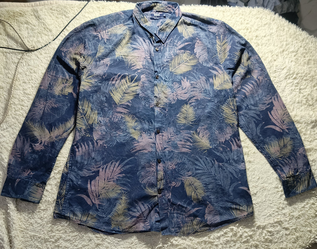 A long-sleeve button-up shirt with a blue tropical palm leaf pattern, laid flat on a white textured blanket. The shirt features shades of navy, medium blue, and light blue-green.