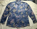 A long-sleeve button-up shirt with a blue tropical palm leaf pattern, laid flat on a white textured blanket. The shirt features shades of navy, medium blue, and light blue-green.