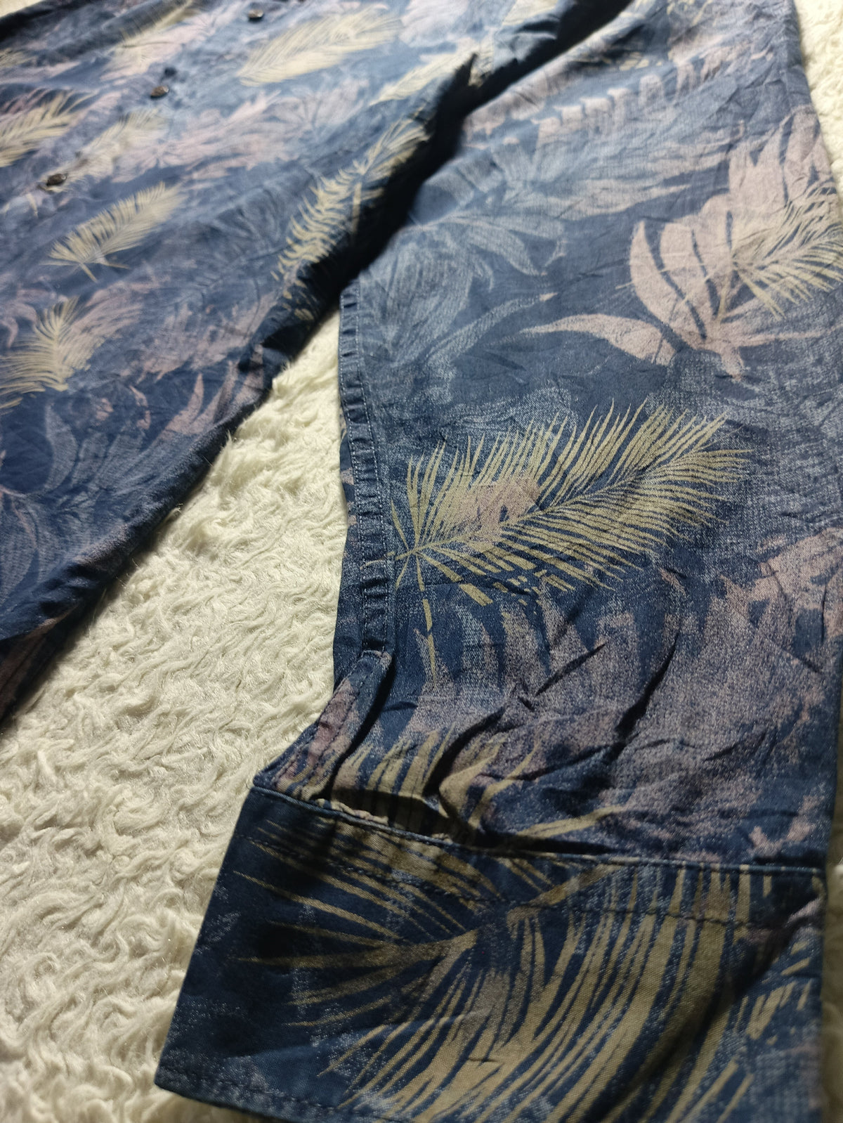 A close-up of a blue tropical leaf print fabric, likely a shirt or towel, laid flat on a textured cream surface. The print features light beige palm leaves over a dark navy blue background.