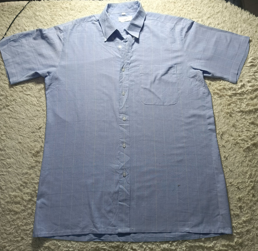 A light blue short-sleeve button-up shirt with a subtle grid pattern, laid flat on a beige shag rug. The shirt features a classic collar, a chest pocket, and a button-down front.